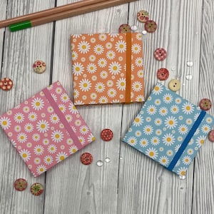 May include: Three small notebooks with daisy patterns in orange, pink, and blue, each with a matching elastic band. Decorative buttons and pencils are scattered around the notebooks on a light wood-grain surface.