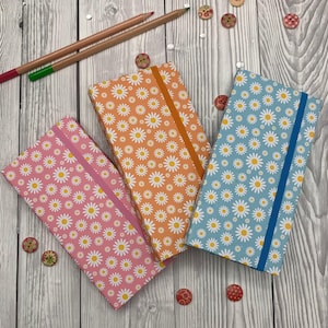 May include: Three notebooks with daisy patterns in pink, orange, and blue, each with a matching elastic band. Two pencils and decorative buttons are scattered around the notebooks on a wooden surface. The notebooks are closed.