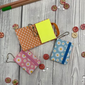 May include: A collection of miniature notebooks and embellishments. One notebook is open, displaying yellow sticky notes. Other notebooks have daisy patterns in pink and blue. Decorative buttons and pencils are scattered around the notebooks.