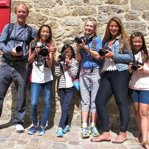 K&ouml;nnte beinhalten: Eine Gruppe von sechs Personen, darunter Erwachsene und Kinder, steht vor einer Steinmauer. Sie halten alle Kameras in der Hand und l&auml;cheln. Die Erwachsenen tragen Freizeitkleidung, w&auml;hrend die Kinder Jeans und T-Shirts tragen. Die Mauer besteht aus grob behauenen Steinen und hat eine rote Holzt&uuml;r im Hintergrund.
