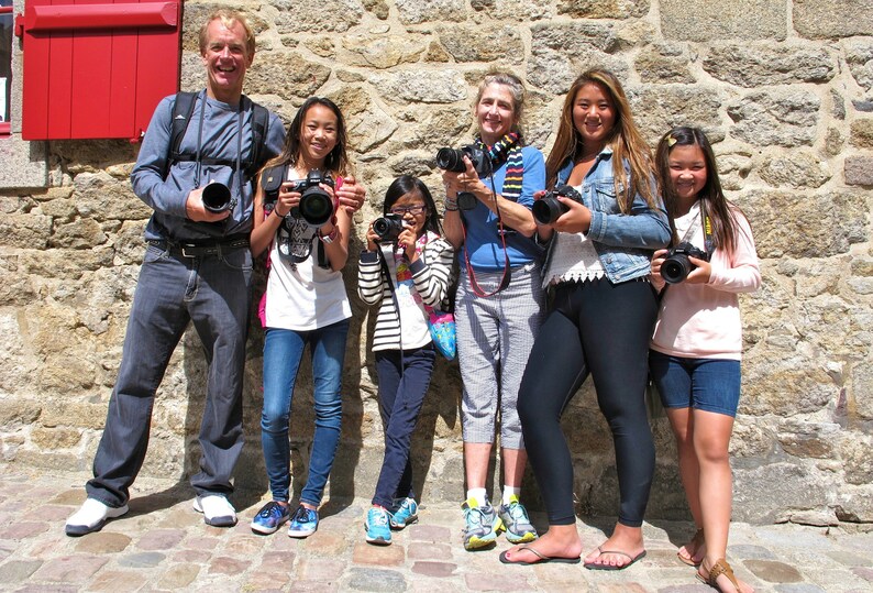 K&ouml;nnte beinhalten: Eine Gruppe von sechs Personen, darunter Erwachsene und Kinder, steht vor einer Steinmauer. Sie halten alle Kameras in der Hand und l&auml;cheln. Die Erwachsenen tragen Freizeitkleidung, w&auml;hrend die Kinder Jeans und T-Shirts tragen. Die Mauer besteht aus grob behauenen Steinen und hat eine rote Holzt&uuml;r im Hintergrund.