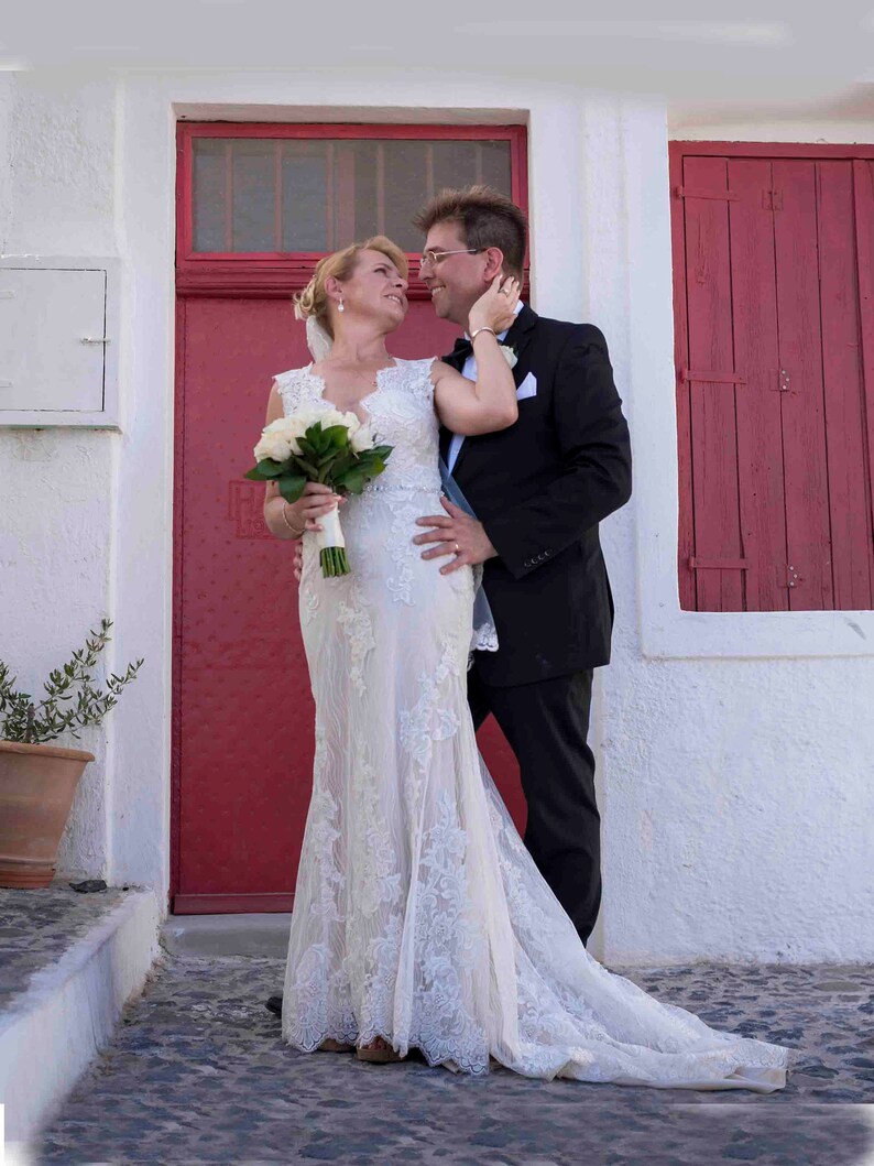 K&ouml;nnte beinhalten: Ein Brautpaar posiert f&uuml;r ein Hochzeitsfoto vor einer roten T&uuml;r. Die Braut tr&auml;gt ein wei&szlig;es Spitzenhochzeitskleid und h&auml;lt einen Strau&szlig; wei&szlig;er Blumen. Der Br&auml;utigam tr&auml;gt einen schwarzen Anzug und ein wei&szlig;es Hemd.