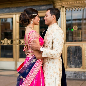 K&ouml;nnte beinhalten: Ein Paar in traditioneller indischer Hochzeitskleidung, die Frau in einem rosa und blauen Lehenga und der Mann in einem wei&szlig;en Sherwani, stehen sich gegen&uuml;ber und l&auml;cheln.