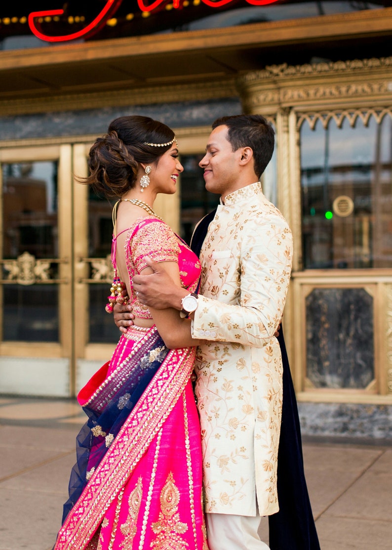 K&ouml;nnte beinhalten: Ein Paar in traditioneller indischer Hochzeitskleidung, die Frau in einem rosa und blauen Lehenga und der Mann in einem wei&szlig;en Sherwani, stehen sich gegen&uuml;ber und l&auml;cheln.