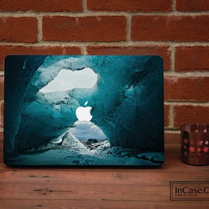 Puede incluir: Una funda para portátil con una impresión fotográfica de alta resolución de una cueva de hielo azul profundo con paredes irregulares y translúcidas, y un logotipo de Apple blanco brillante centrado en medio de la escena gélida.