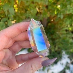 May include: A clear, iridescent crystal point with a prism effect, held against a blurred green background. The crystal reflects a spectrum of colors, including blues, yellows, and oranges. The crystal is approximately 4 inches long.