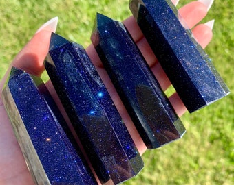 May include: Four blue and black star-shaped gemstone points, also known as blue goldstone, are arranged in a row on a hand. The points are polished and have a sparkly, glittery appearance.