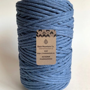 May include: A large spool of blue-grey macrame cord. The cord is tightly wound and has a textured appearance. A small brown tag with the text "Rock Mountain Co." and contact information is attached to the spool.