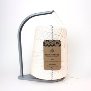 May include: A white cotton twine spool on a gray metal stand. The spool has a brown paper tag with the text "Rock Mountain Co. info@rockmountain.co SHOP https://rockmountain.co INSTAGRAM @rockmountainco"