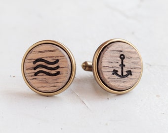 May include: A pair of wooden cufflinks with a brass finish. One cufflink features a wave design, while the other features an anchor design.