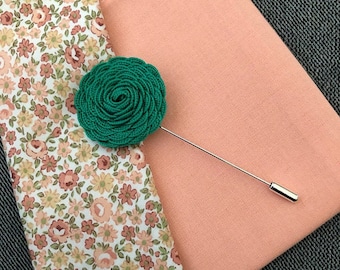 May include: A green fabric rose boutonniere with a silver pin on a peach colored fabric background.