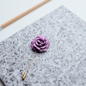 May include: A purple velvet rose lapel pin with a gold stem. The pin is on a gray textured surface.