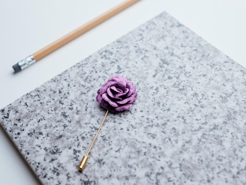 May include: A purple velvet rose lapel pin with a gold stem. The pin is on a gray textured surface.