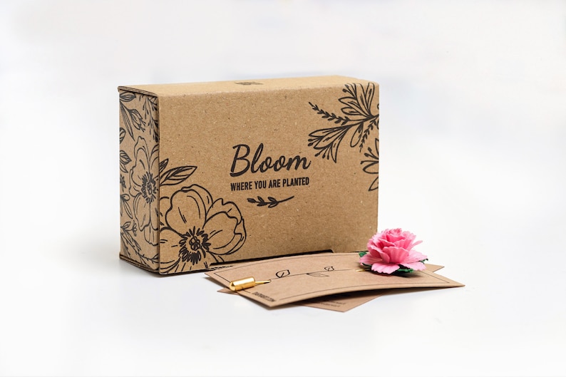 May include: A brown cardboard box with a floral design and the text "Bloom Where You Are Planted". A pink carnation flower is on top of a brown card.