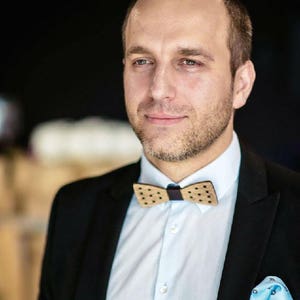 May include: A man wearing a black suit jacket with a light blue shirt, a brown and white polka dot bow tie, and a light blue pocket square.