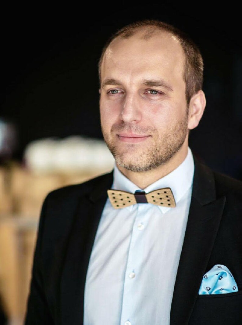 May include: A man wearing a black suit jacket with a light blue shirt, a brown and white polka dot bow tie, and a light blue pocket square.