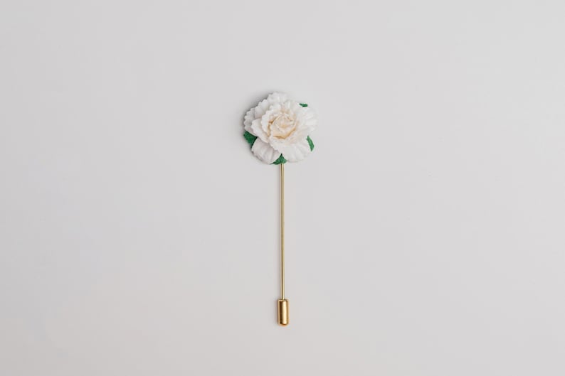 May include: A white flower lapel pin with a gold stem and a gold pin on a white background.