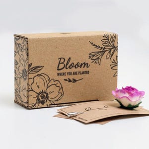 A cardboard box with a floral design and the text "Bloom" printed on it, along with a small pink rose-shaped object and some brown paper cards.
