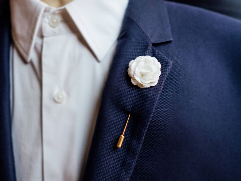 May include: A white fabric flower pinned to a navy blue suit jacket with a gold pin.