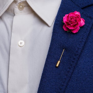 May include: A dark blue blazer with a pink rose boutonniere and a gold pin.