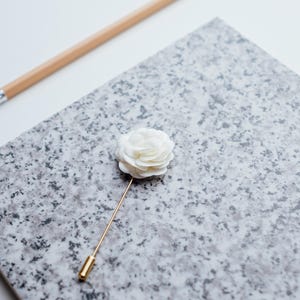 May include: A white fabric flower lapel pin with a gold stem. The pin is on a gray and white speckled surface.