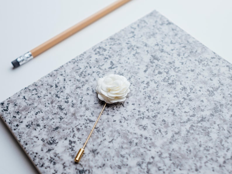 May include: A white fabric flower lapel pin with a gold stem. The pin is on a gray and white speckled surface.