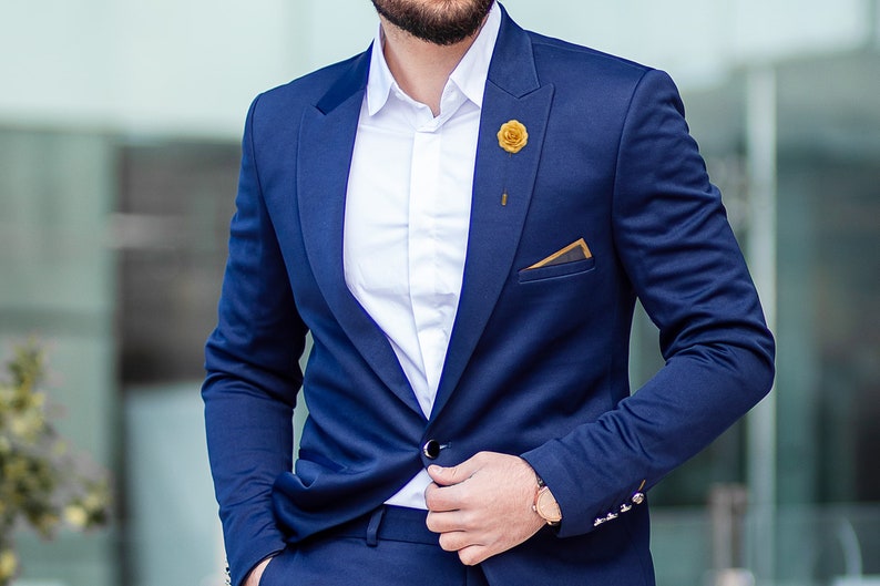 May include: A navy blue suit jacket with a white button-down shirt underneath. The jacket has a gold flower lapel pin and a pocket square with a gold trim.