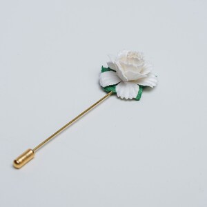 May include: A white flower with green leaves on a gold pin.