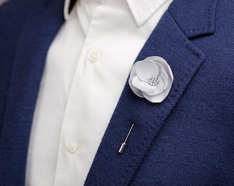 May include: A white flower lapel pin with a silver pin backing is attached to a navy blue blazer. The flower has a cluster of small, sparkling embellishments in the centre. A white shirt is visible beneath the blazer.