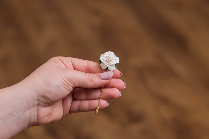 May include: A white flower with green leaves on a gold pin. The pin is held in a hand.