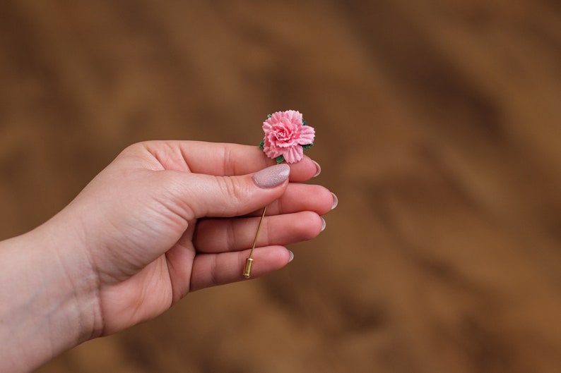 May include: A pink carnation flower on a gold pin. The pin is held in a hand.