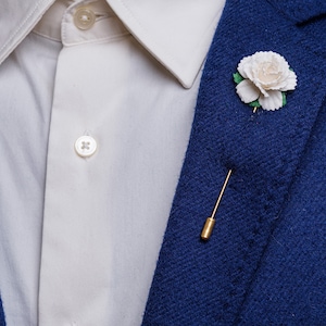 May include: A white flower lapel pin with green leaves, attached to a gold pin, on a blue blazer.
