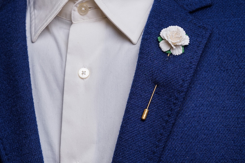 May include: A white flower lapel pin with green leaves, attached to a gold pin, on a blue blazer.