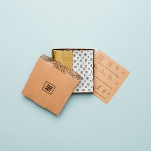 May include: A brown cardboard box with the word "The Way" printed on it. The box is open and a smaller box is inside. The smaller box is white with a blue and white patterned fabric inside. A brown paper with instructions is next to the boxes.