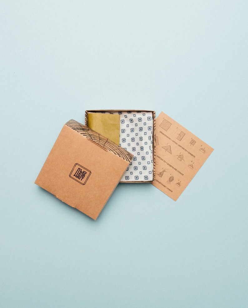 May include: A brown cardboard box with the word "The Way" printed on it. The box is open and a smaller box is inside. The smaller box is white with a blue and white patterned fabric inside. A brown paper with instructions is next to the boxes.