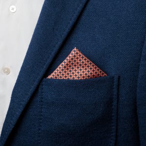 May include: Close-up of a navy blue blazer with a peach-colored pocket square. The pocket square features a geometric pattern. A white dress shirt is visible beneath the blazer.