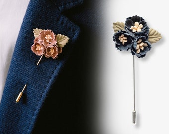 Può includere: Un paio di spille da bavero con un gruppo di tre fiori in tessuto. Una spilla ha fiori rosa con foglie verdi e una spilla dorata. L'altra spilla ha fiori blu con centri pesca e foglie beige e una spilla argentata.