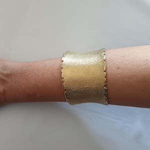 May include: A wide, gold-colored cuff bracelet with a textured surface and a scalloped edge. The bracelet is worn on a person's arm, showcasing its metallic sheen and unique design. The cuff is approximately 2 inches wide.