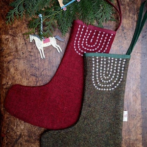 Könnte beinhalten: Zwei Weihnachtsstrümpfe in Burgunderrot und Olivgrün mit weißen, gestickten Sternmustern. Der burgunderfarbene Strumpf hat eine burgunderfarbene Schlaufe, und der olivgrüne Strumpf hat eine grüne Schlaufe. Holzornamente eines Hundes auf einem Skateboard und eines Pferdes liegen obenauf.