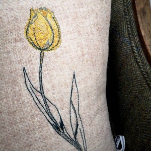 Op de afbeelding: Een beige kussen met een geborduurd geel tulpenmotief. Het kussen wordt getoond tegen een groene en bruine tweed stof.