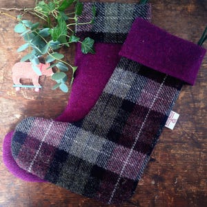 May include: A purple and plaid Christmas stocking with a green loop at the top. The stocking is made of wool and has a plaid pattern with shades of purple, black, and gray. The stocking is lying on a wooden surface.