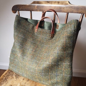 May include: A large green and brown tweed tote bag with leather handles and brass accents. The bag is sitting on a wooden chair.