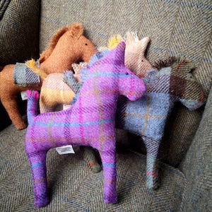 May include: Five plush toy horses made of tweed fabric in various colors, including purple, brown, yellow, and blue. The horses are sitting on a tweed armchair.