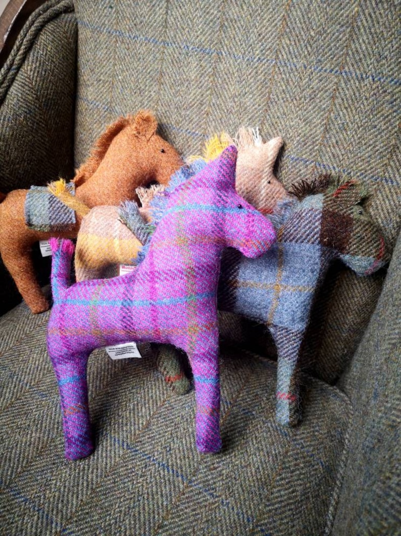 May include: Five plush toy horses made of tweed fabric in various colors, including purple, brown, yellow, and blue. The horses are sitting on a tweed armchair.