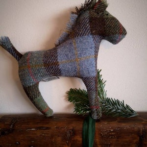 May include: A blue, green, and brown plaid horse toy made of tweed fabric. The horse is standing on a wooden surface with green foliage behind it.