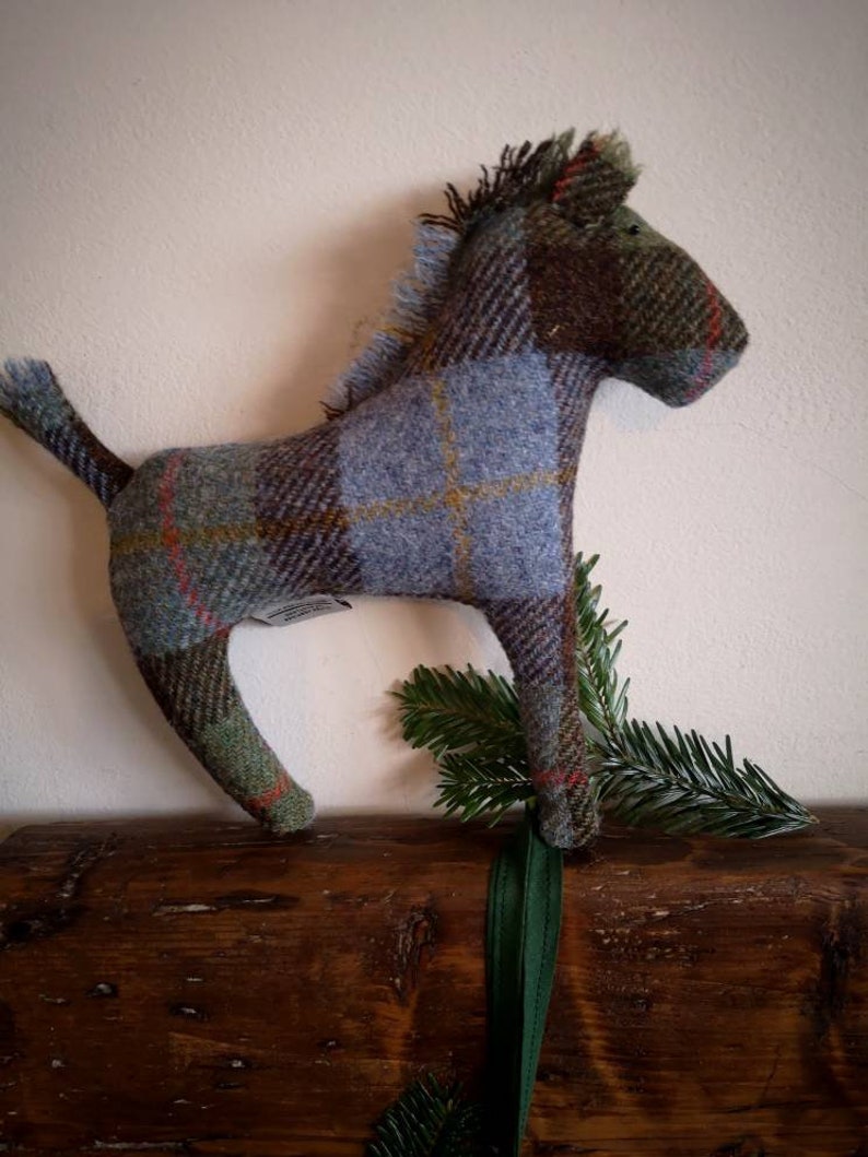 May include: A blue, green, and brown plaid horse toy made of tweed fabric. The horse is standing on a wooden surface with green foliage behind it.