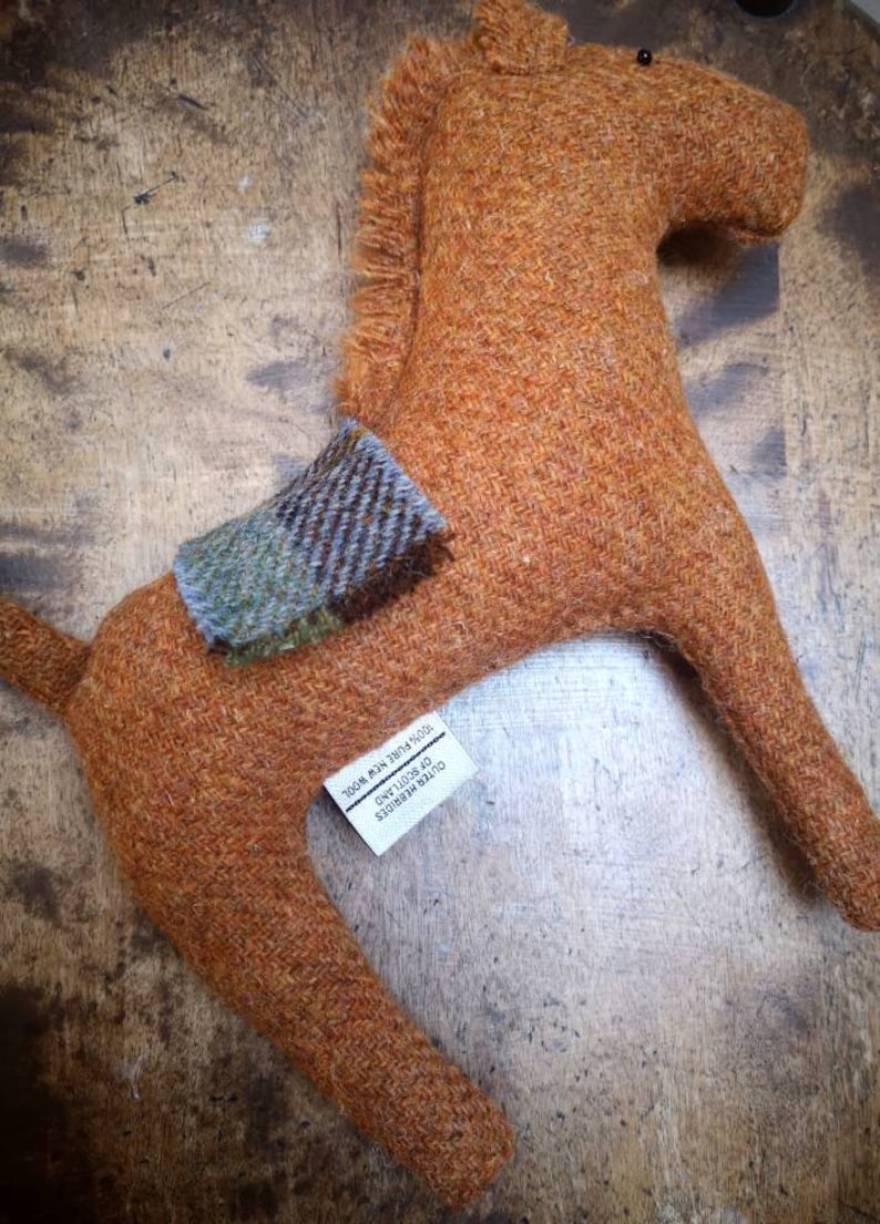 May include: A brown wool horse toy with a plaid saddle. The toy is handmade and has a tag that reads "OUTER HEBRIDES OF SCOTLAND  MADE IN THE UK  2808 MIN 780741205".