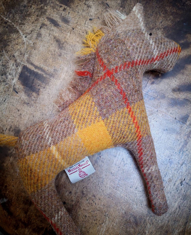 May include: A brown and yellow plaid horse toy with red accents. The toy is made of wool and has a label that says "Harris Tweed" on it.