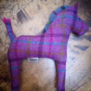 May include: A pink and blue plaid horse stuffed animal with a long tail. The horse is made of tweed and has a label that reads "100% Pure New Wool, Made in Scotland".