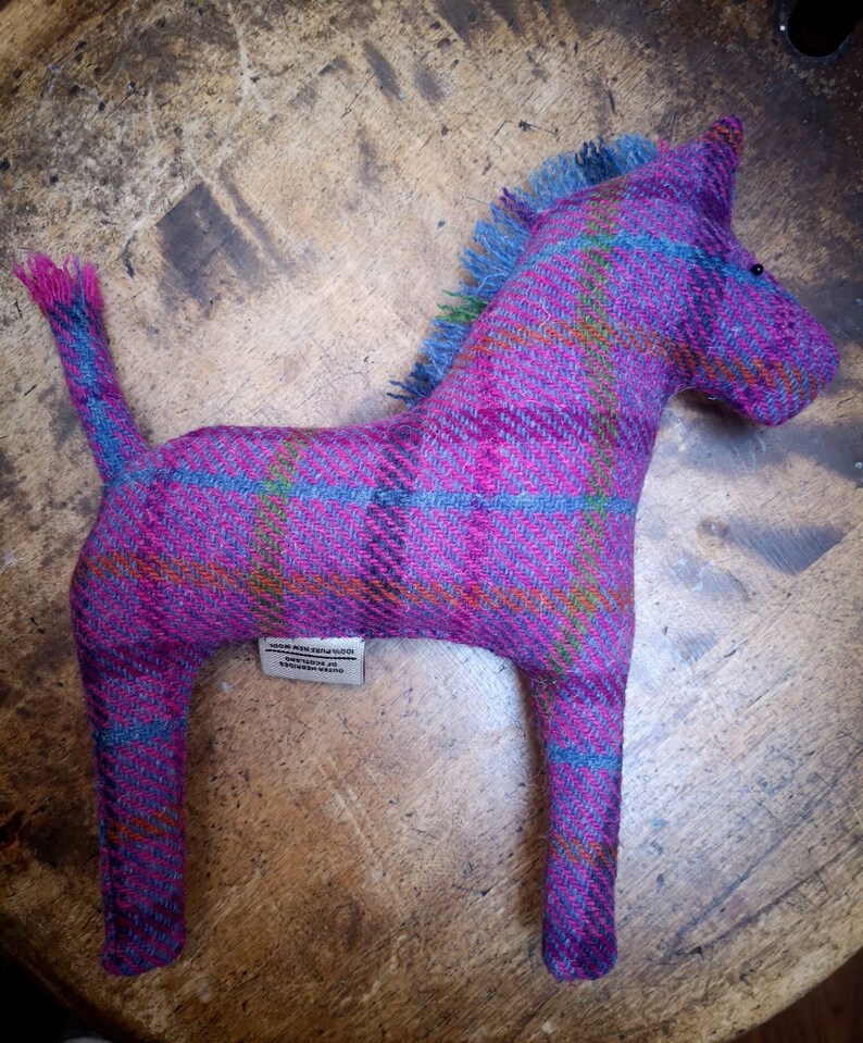 May include: A pink and blue plaid horse stuffed animal with a long tail. The horse is made of tweed and has a label that reads "100% Pure New Wool, Made in Scotland".